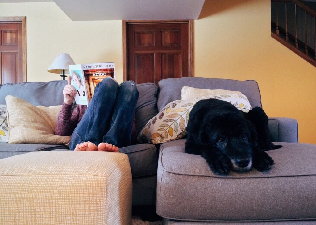 Mujer tumbada en un sofá leyendo una revista y perro negro tumbado a su lado